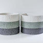 A textured ceramic pot, displaying stacked bands of white, green, and gray, sits on a smooth white surface against a plain white wall.