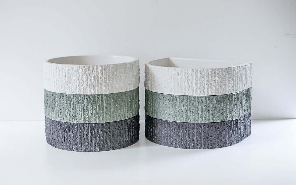 A textured ceramic pot, displaying stacked bands of white, green, and gray, sits on a smooth white surface against a plain white wall.