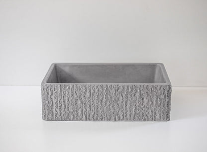 A rectangular, textured concrete planter with a ribbed surface rests on a smooth, white surface against a plain white background, creating a minimalist aesthetic.