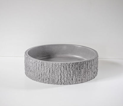 A round, textured gray concrete bowl sits on a white surface, surrounded by plain white walls, showcasing its modern, minimalist design.