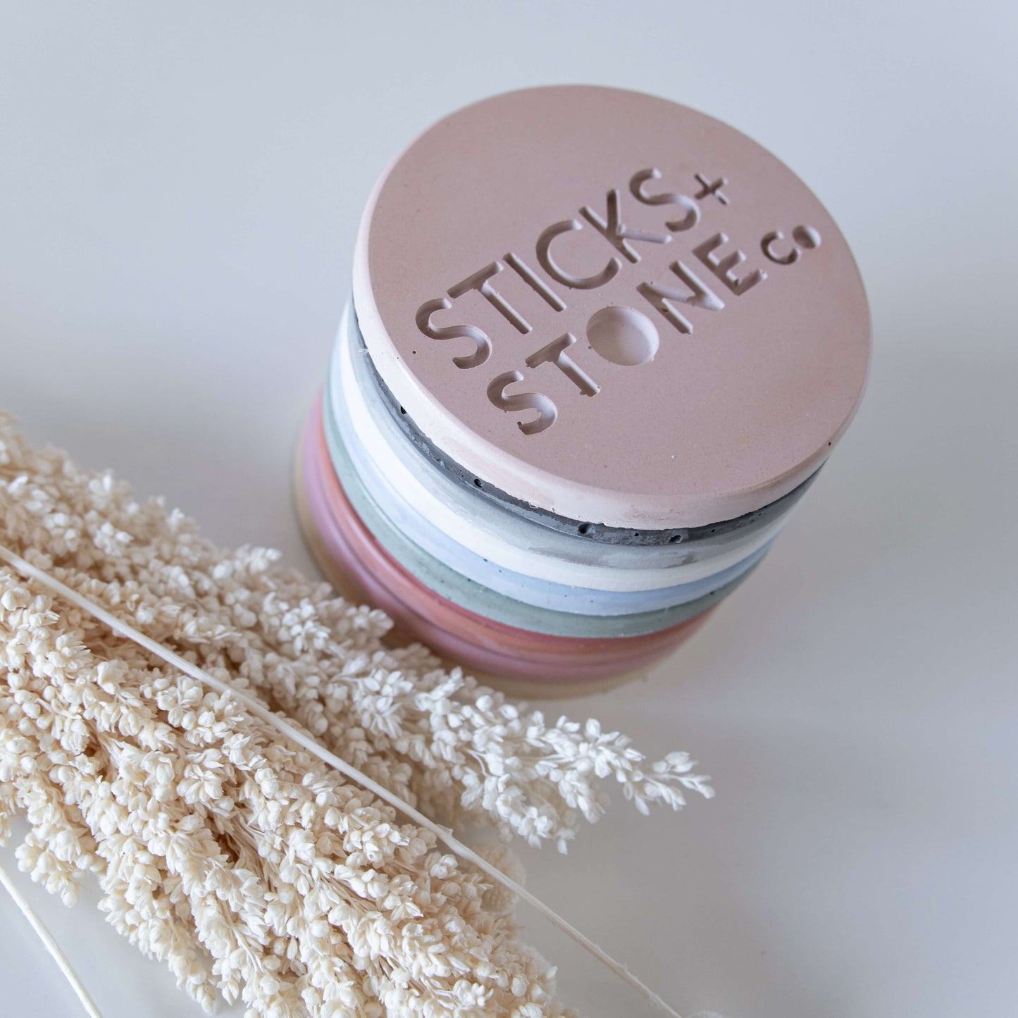 A stack of colorful, round coasters embossed with "STICKS + STONE co." rests on a white surface beside dried beige flowers, creating a minimalist, natural decor setting.