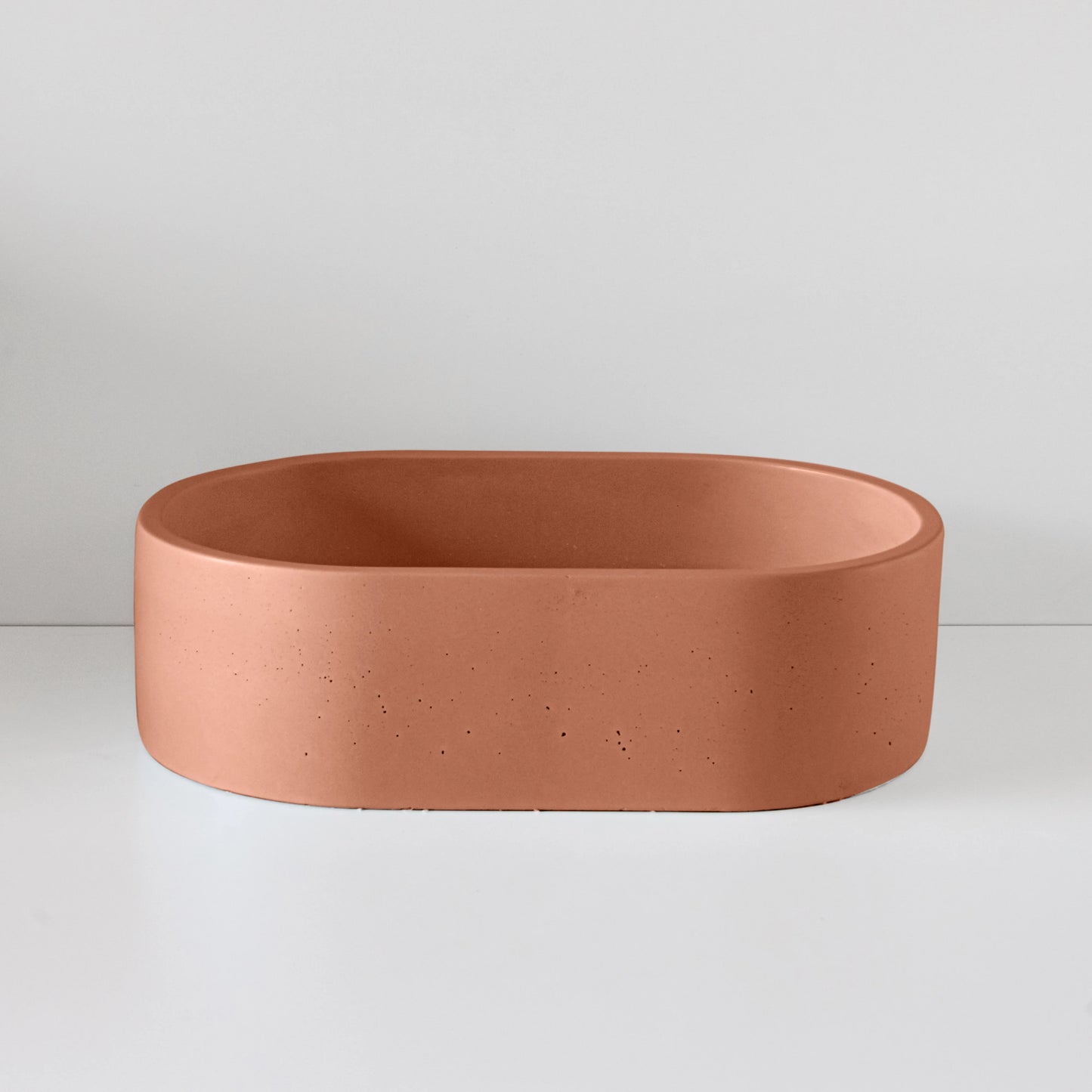 Oval, terracotta-colored basin sits stationary on a plain white surface, with a smooth, minimalist design and small speckled texture under soft lighting.