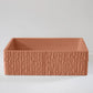 A terracotta-colored rectangular planter with a textured exterior sits on a smooth, light gray surface in a minimalistic setting.