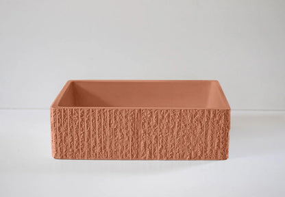 A terracotta-colored rectangular planter with a textured exterior sits on a smooth, light gray surface in a minimalistic setting.