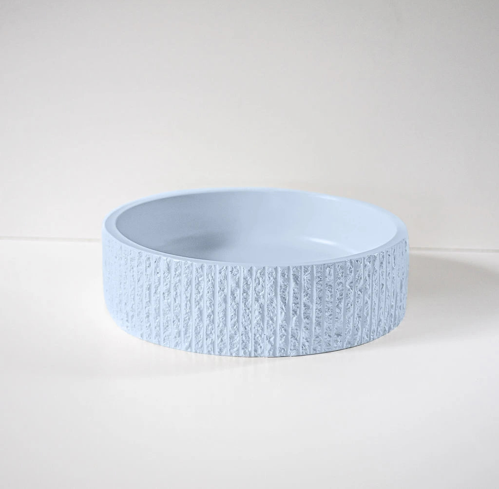 A light blue, textured ceramic bowl sits on a smooth, white surface against a pale background. The bowl features vertical ridges around its exterior, providing a tactile appearance.