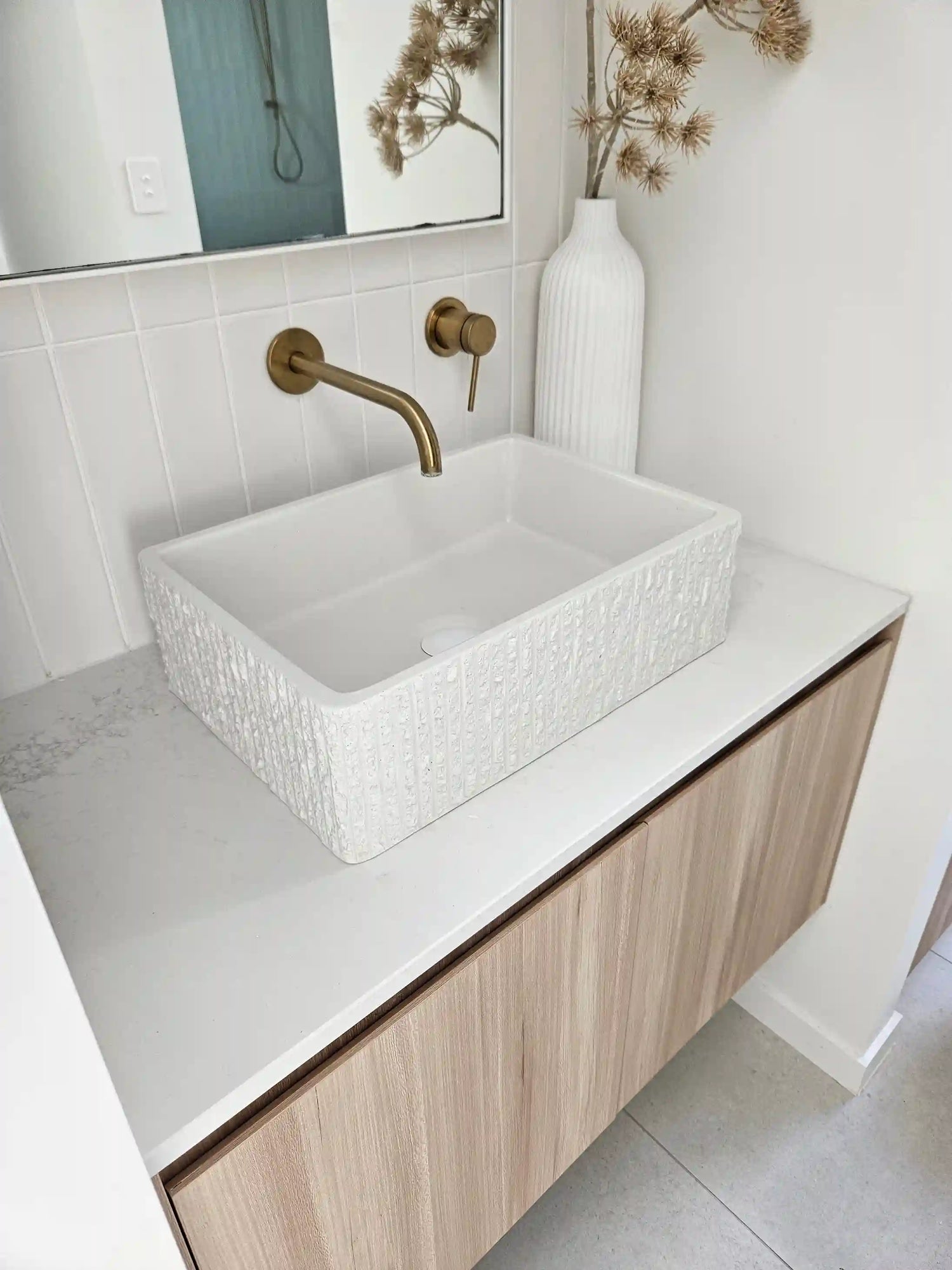 Modern bathroom vanity with a textured concrete basin, brass wall-mounted faucet, and decorative dried plants in a white vase, set against a white tiled wall.