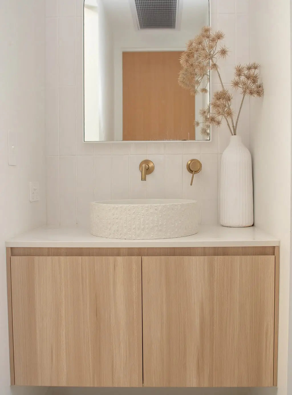 Modern bathroom featuring a white concrete round rugged basin on a wooden vanity with gold tapware, accompanied by a decorative vase and dried flowers, enhancing the minimalist aesthetic.