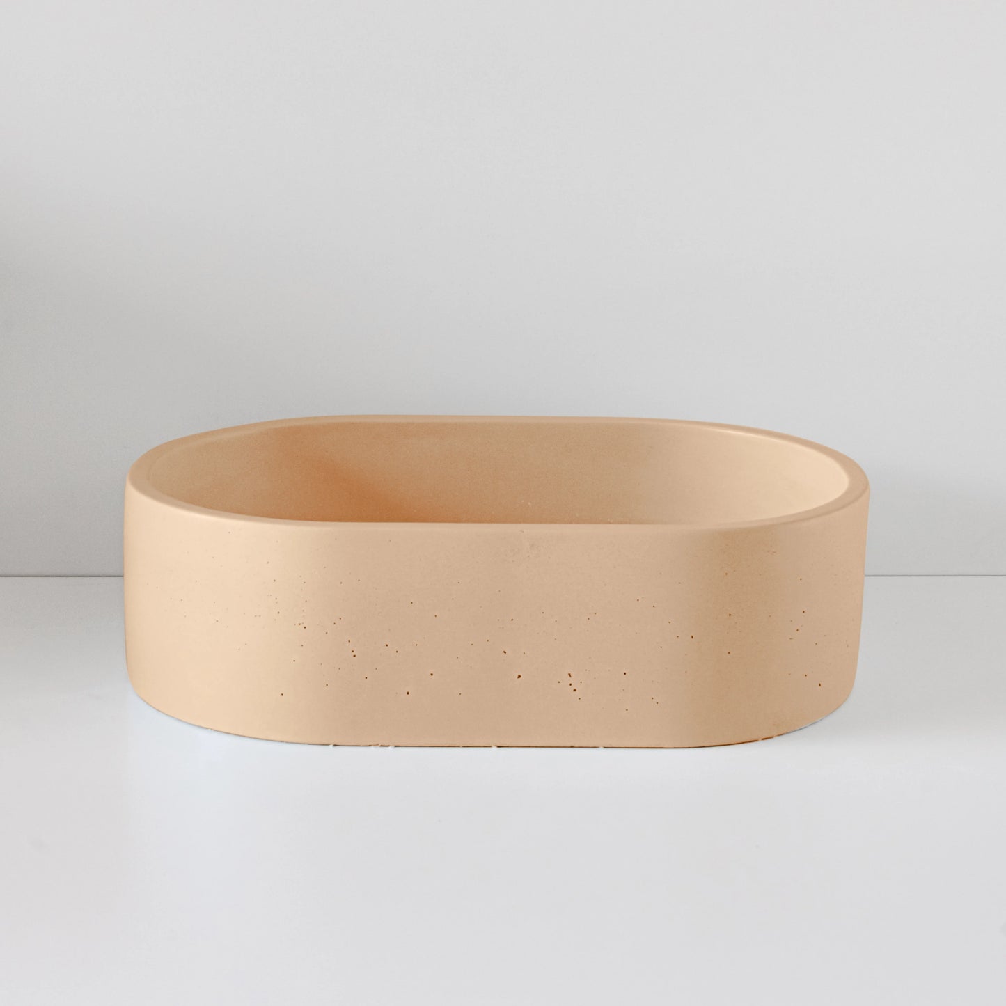 A minimalist, oval-shaped, beige vessel sits empty on a smooth, white surface, set against a plain, light-colored background.