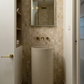 A minimalist bathroom featuring a cylindrical concrete basin vanity with brass tapware, an arched mirror, and beige square-tiled walls.