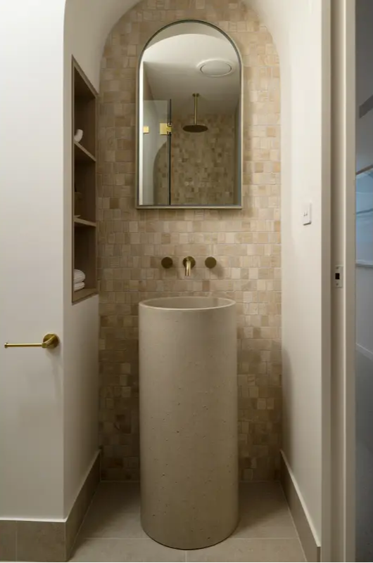 A minimalist bathroom featuring a cylindrical concrete basin vanity with brass tapware, an arched mirror, and beige square-tiled walls.