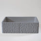 A square-shaped, gray stone basin with textured, vertical stripes sits on a smooth, light-colored surface against a plain background.