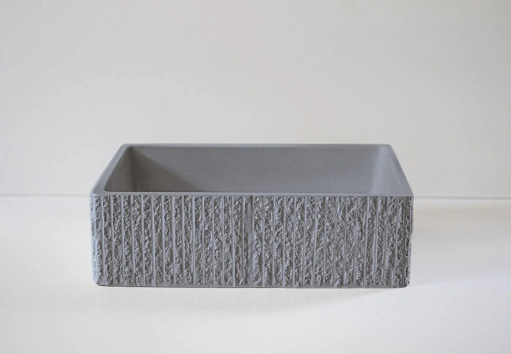 A square-shaped, gray stone basin with textured, vertical stripes sits on a smooth, light-colored surface against a plain background.