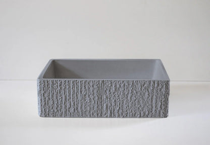 A square-shaped, gray stone basin with textured, vertical stripes sits on a smooth, light-colored surface against a plain background.