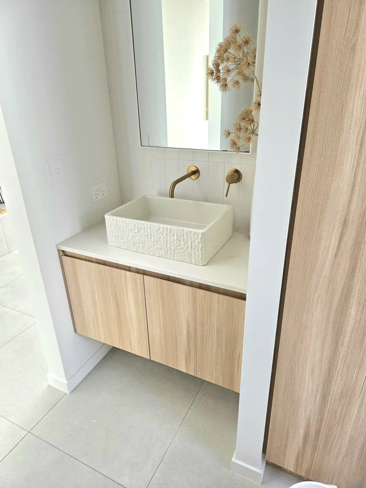 Bathroom vanity with light wood cabinetry, featuring a rugged concrete rectangle basin and brass wall-mounted faucet with white tiled backsplash.