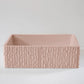 A rectangular, textured pink ceramic basin sits empty on a smooth, light surface, with a plain, neutral-colored background.