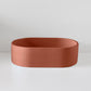 A terracotta-colored, oval-shaped basin sits on a white surface against a plain, light gray background.