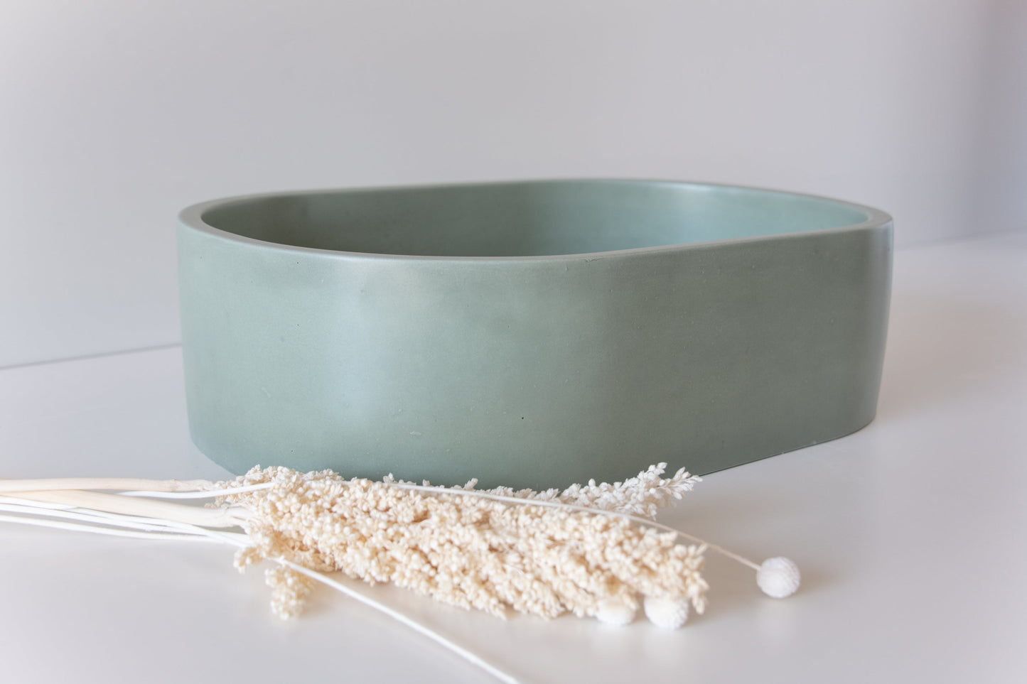 A smooth, green, oval ceramic bowl sits on a white surface, accompanied by a cluster of dried, white flowers placed in front of it.